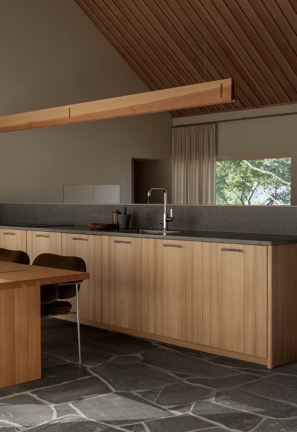 Warm, minimalist kitchen-diner with wooden units, granite worktop, exposed timber ceiling, flagstone floor and bespoke wooden dining table | Home tour - a contemporary take on a traditional Swedish longhouse in Österlen, Skåne | These Four Walls blog